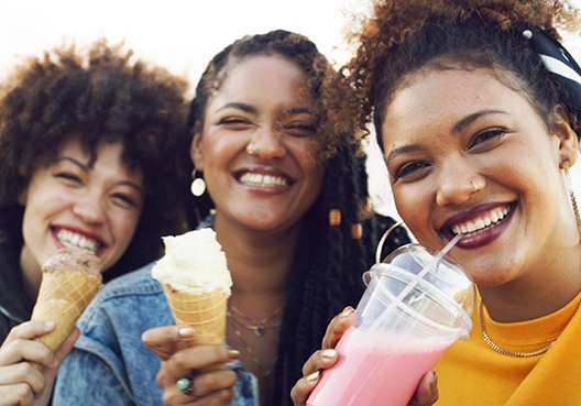 Three young women eating ice cream and drinking milkshakes looking and laughing at the camera